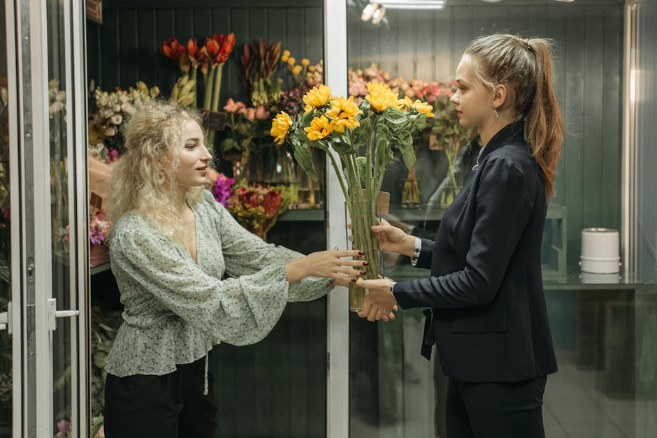 A florist with light brown hair tied back in a high ponytail, dressed in a dark blazer, is handing a tall, clear glass vase filled with bright yellow sunflowers to a woman with curly blonde hair weari