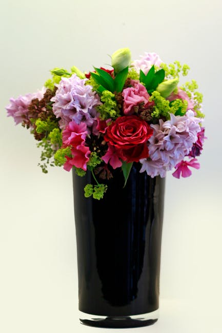 A vibrant floral arrangement in a tall, sleek black vase, featuring a mix of soft pink, lavender, white, and bright red flowers. The bouquet includes roses, hydrangeas, and lisianthus, with greenery a