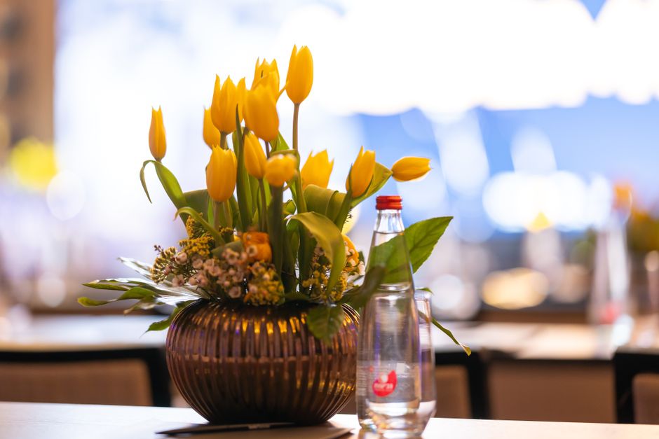 A floral arrangement featuring tall yellow tulips with closed buds and bright green leaves, placed in a dark amber ribbed glass vase. Surrounding the tulips are small clusters of delicate pink and whi