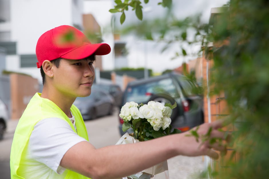 A delivery person wearing a red cap and a high-visibility yellow vest is holding a bouquet of white hydrangeas wrapped in clear plastic. They are positioned outdoors on a street lined with parked cars