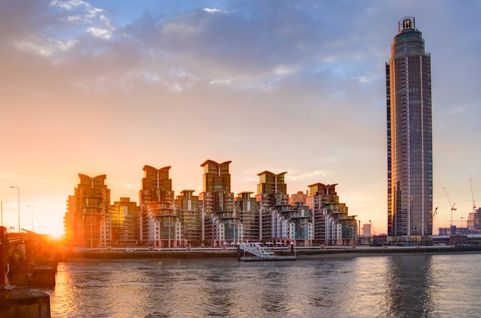A cityscape featuring a collection of contemporary residential buildings with distinctive, staggered rooftop terraces, illuminated by the warm glow of a setting sun. A tall, cylindrical skyscraper wit