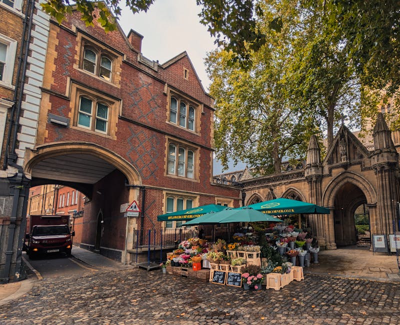 A vibrant outdoor flower stall situated on a cobbled street near an historic Gothic-style stone archway and red-brick building with arched windows. The stall, shaded by a large green umbrella bearing 